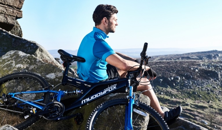 Man sat on a rock staring into the distance next to a blue and black Muddy Fox mountain bike.